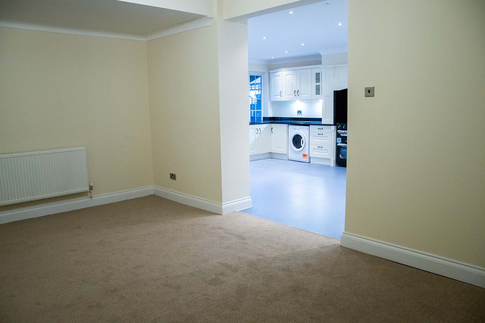 Empty carpeted room leading into the brightly-lit kitchen